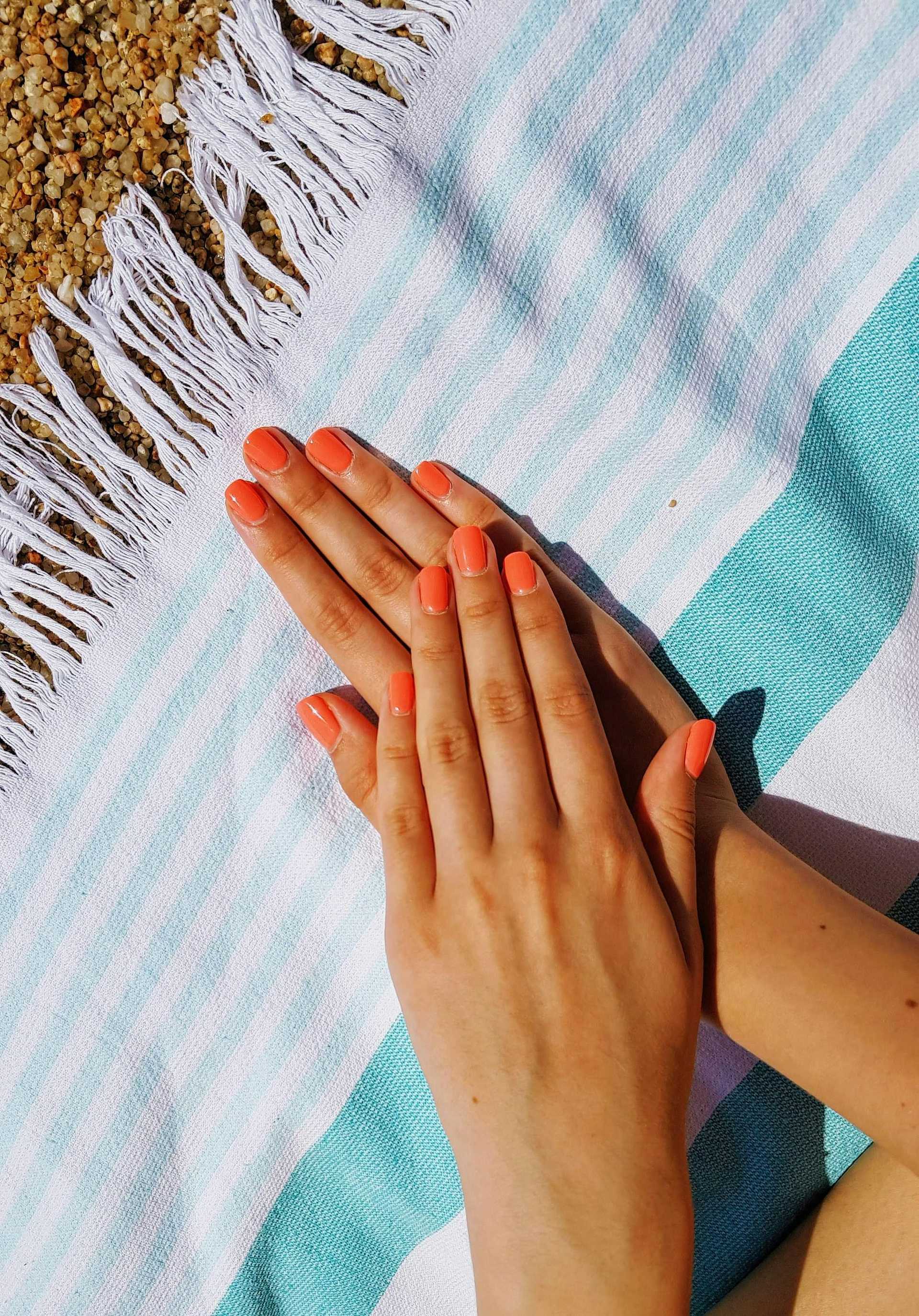 Hands with coral nail polish resting on a striped beach towel near pebbles.